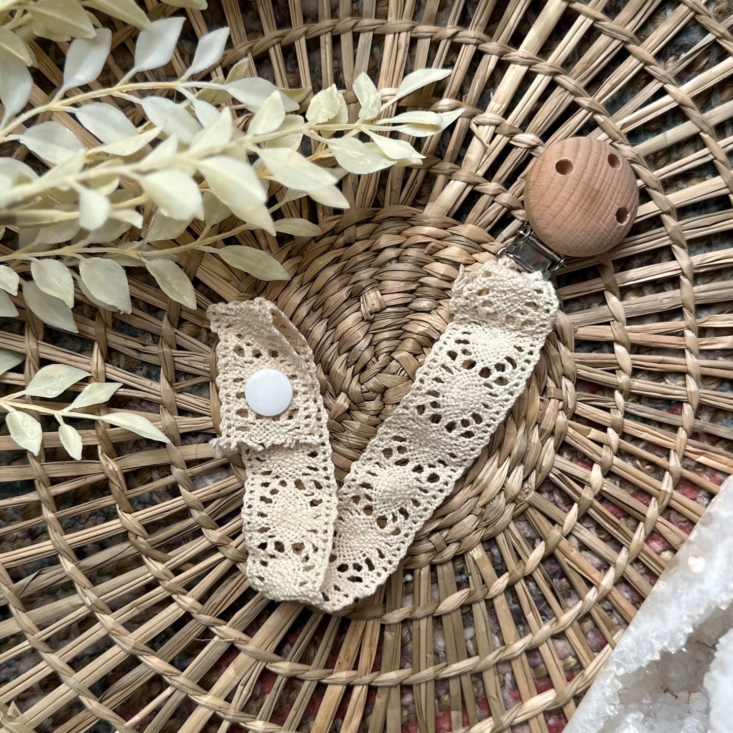 A cream lace pacifier holder with a wooden clip and white lace detailing, displayed on a woven mat.